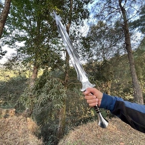 Puede incluir: Una espada plateada con una hoja puntiaguda y un mango ornamentado. La espada se sostiene contra un tel&oacute;n de fondo de &aacute;rboles y follaje, lo que sugiere un entorno al aire libre. El mango presenta un agarre marr&oacute;n oscuro y detalles plateados.