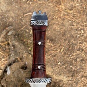 May include: A close-up of a handcrafted knife handle. The handle is made of dark brown wood with silver accents. The top of the handle has a silver cap with a decorative design. The blade is visible at the bottom.