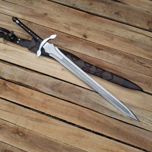 Peut inclure: Une épée en métal argenté dotée d'une longue lame à double tranchant avec un motif rainuré central. L'épée possède un manche noir avec des rivets argentés et une garde incurvée, accompagnée d'un fourreau en cuir marron foncé texturé.