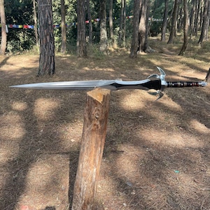 May include: A silver sword with a black handle and a decorative design on the pommel. The sword is resting on a wooden log in a forest setting.
