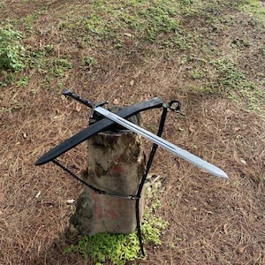 May include: A silver sword with a black handle and a black sheath. The sword is resting on a weathered wooden stump, with a black stand supporting the sheath. The background is a mix of brown and green foliage.