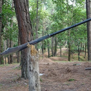May include: A long, black-handled sword with a curved blade is propped against a tree trunk in a forest setting.