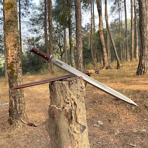 May include: A silver sword with a brown leather-wrapped handle and sheath is displayed on a tree stump. The sword has a wide, flat blade and a guard. The background shows a forest with tall trees.