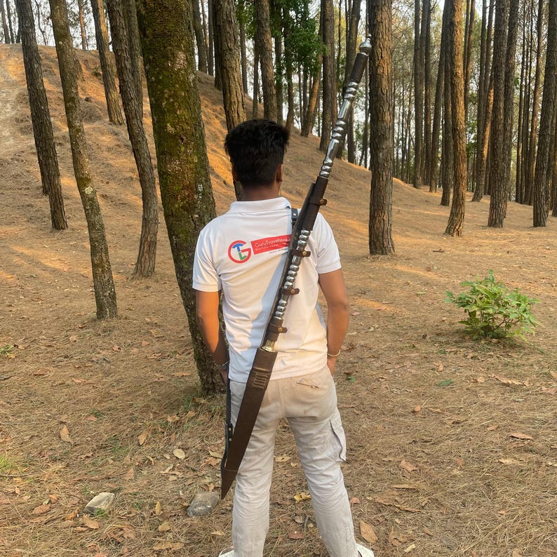 May include: A person wearing a white shirt with a red logo is carrying a long, silver-colored sword in a brown leather scabbard on their back. The sword has a decorative handle and is strapped to the person's shoulder with a brown leather strap. The person is standing in a forest setting.