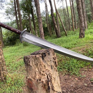 May include: A large, silver sword with a brown handle and a decorative silver end cap rests on a tree stump. The sword has a wide blade and a curved tip. The background features a forest with tall trees and green grass.