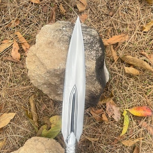 May include: A silver spearhead with a black and silver design. The spearhead is pointed and has a sharp edge. It is resting on a rock.