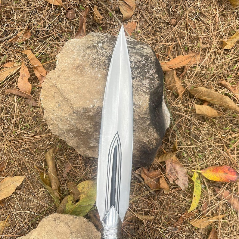 May include: A silver spearhead with a black and silver design. The spearhead is pointed and has a sharp edge. It is resting on a rock.