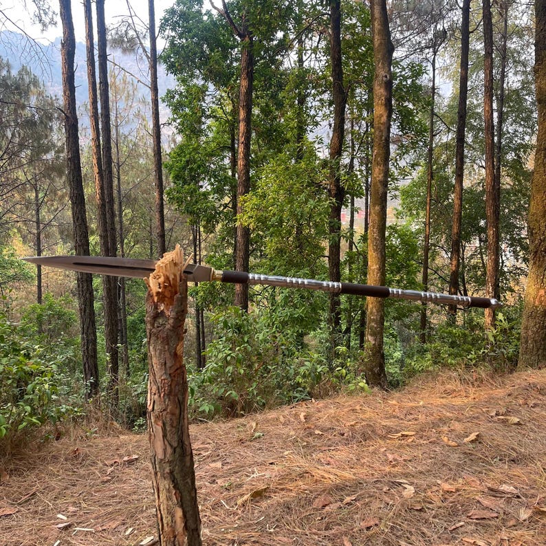 May include: A silver and black spear with a wooden handle is propped against a tree trunk in a forest setting.