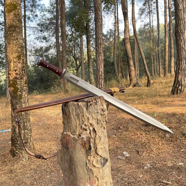 May include: A silver sword with a brown leather-wrapped handle and a matching sheath rests on a tree stump. The sword has a wide, flat blade and a decorative hilt. The background features a forest setting.
