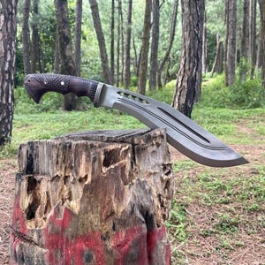Könnte beinhalten: Ein silberfarbenes Messer mit einem Holzgriff liegt auf einem verwitterten Baumstumpf in einer Waldlandschaft. Das Messer hat eine gebogene Klinge mit einer scharfen Spitze und einer gezackten Kante.