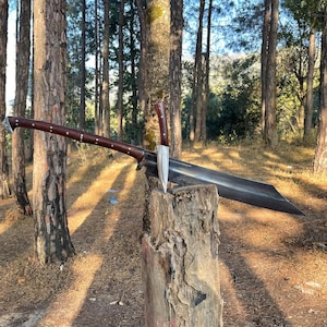 Puede incluir: Una espada grande, curva, negra con un mango de madera marrón está apoyada en un tocón de árbol en un entorno boscoso. La espada tiene un cuchillo más pequeño, plateado, unido al mango.
