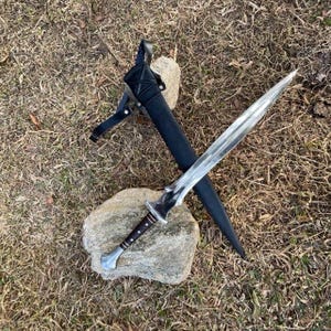 Puede incluir: Una espada plateada y negra con una funda de cuero. La espada tiene un mango detallado y una hoja afilada y puntiaguda. La funda es negra y tiene una correa. La espada y la funda descansan sobre una piedra.