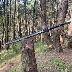 May include: A long, silver sword with a black handle and a silver pommel. The sword is lying on a tree stump in a forest setting.