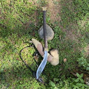 May include: A curved blade sword with a brown leather-wrapped handle and gold accents. The sword is sheathed in a patterned leather scabbard with a black strap. The sword rests on a bed of green grass and small rocks.