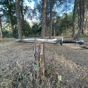 Puede incluir: Una espada larga y plateada con una hoja puntiaguda y un mango adornado, apoyada sobre un toc&oacute;n de &aacute;rbol en un entorno forestal. El mango presenta detalles en negro y plata. La espada es un arma de estilo medieval.