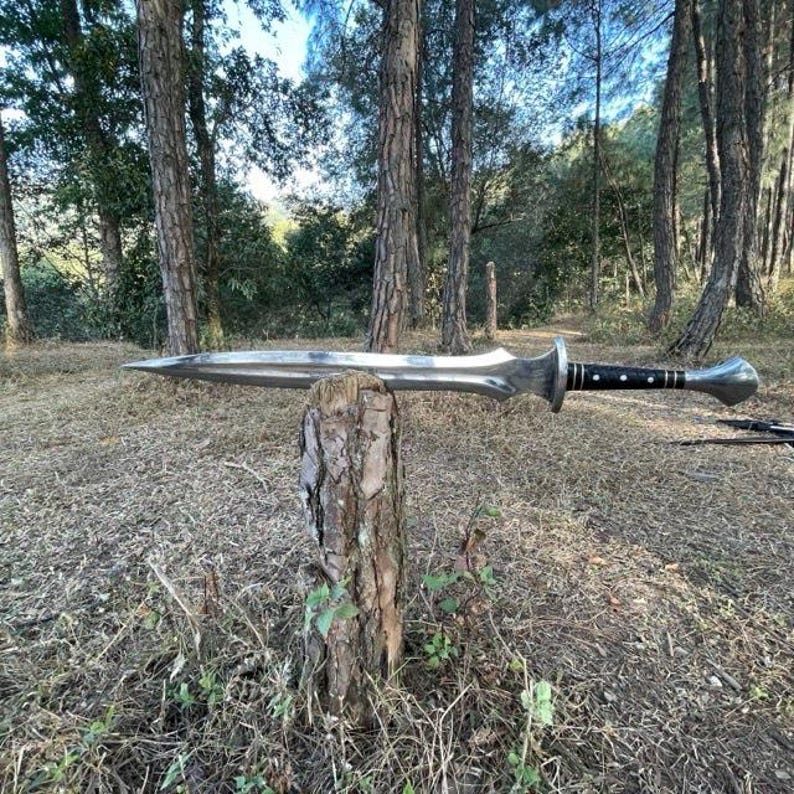 Puede incluir: Una espada larga y plateada con una hoja puntiaguda y un mango adornado, apoyada sobre un toc&oacute;n de &aacute;rbol en un entorno forestal. El mango presenta detalles en negro y plata. La espada es un arma de estilo medieval.