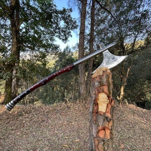 May include: A silver axe with a wooden handle, the axe is stuck in a tree stump in a forest setting.