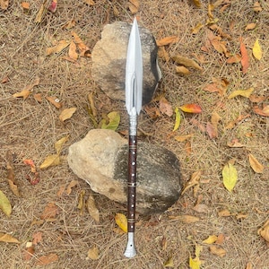 May include: A silver spear with a wooden handle and decorative metal accents. The spear is lying on a bed of dry leaves and rocks.