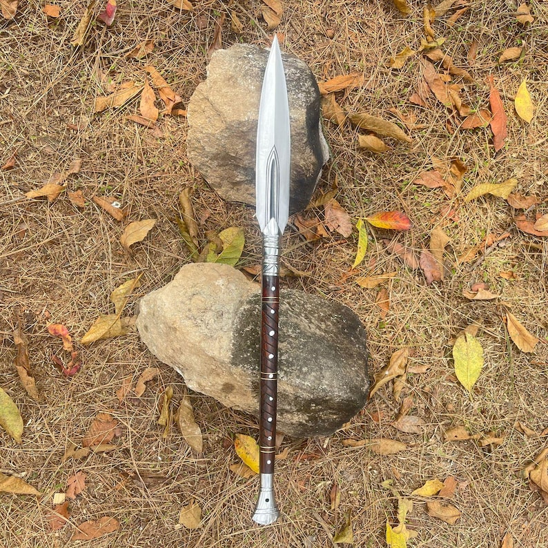 May include: A silver spear with a wooden handle and decorative metal accents. The spear is lying on a bed of dry leaves and rocks.