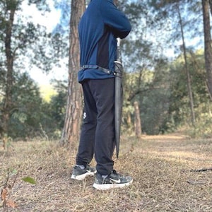 Puede incluir: Una espada larga de color gris oscuro con empu&ntilde;adura y vaina negras. La espada se sujeta verticalmente, sujeta a un cintur&oacute;n negro. La persona lleva una chaqueta azul, pantalones negros y zapatillas deportivas negras y blancas. El fondo es un bosque.