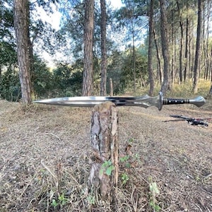 Puede incluir: Una espada plateada con una hoja puntiaguda y una empu&ntilde;adura ornamentada descansa sobre un toc&oacute;n de &aacute;rbol. La espada tiene un mango negro y una guarda decorativa. El fondo presenta un entorno forestal con &aacute;rboles altos y hojas secas.