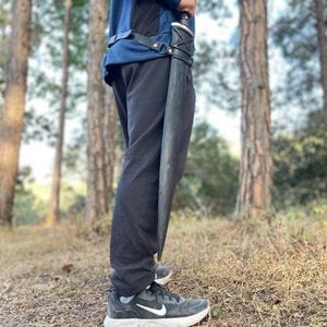 Puede incluir: Una espada negra en una vaina negra se sostiene verticalmente. La espada est&aacute; sujeta a un cintur&oacute;n de cuero negro. La persona lleva pantalones negros y zapatillas negras y grises. El fondo es un bosque con &aacute;rboles altos.
