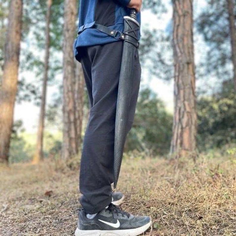 Puede incluir: Una espada negra en una vaina negra se sostiene verticalmente. La espada est&aacute; sujeta a un cintur&oacute;n de cuero negro. La persona lleva pantalones negros y zapatillas negras y grises. El fondo es un bosque con &aacute;rboles altos.