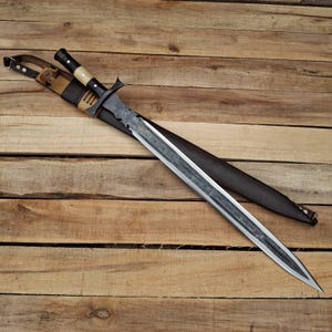 May include: A double-edged sword with a dark, patterned steel blade featuring intricate engravings. The handle has a black grip with a light-colored accent and a metal crossguard. It is partially inserted into a dark brown leather scabbard.