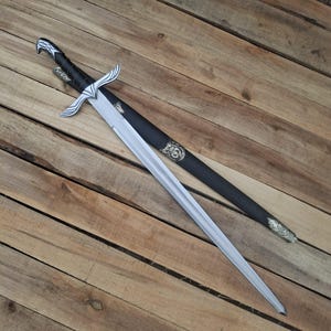 May include: A silver-colored metal sword with a pointed blade and a black wrapped handle. The crossguard features wing-like silver accents. It is accompanied by a black scabbard with ornate silver-colored metal fittings at the top and bottom tip.