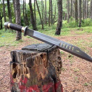 May include: A gray metal sword with a brown and black handle rests on a tree stump. The sword has a long, flat blade with several cut-out holes. The handle is wrapped in a textured material with gold accents.