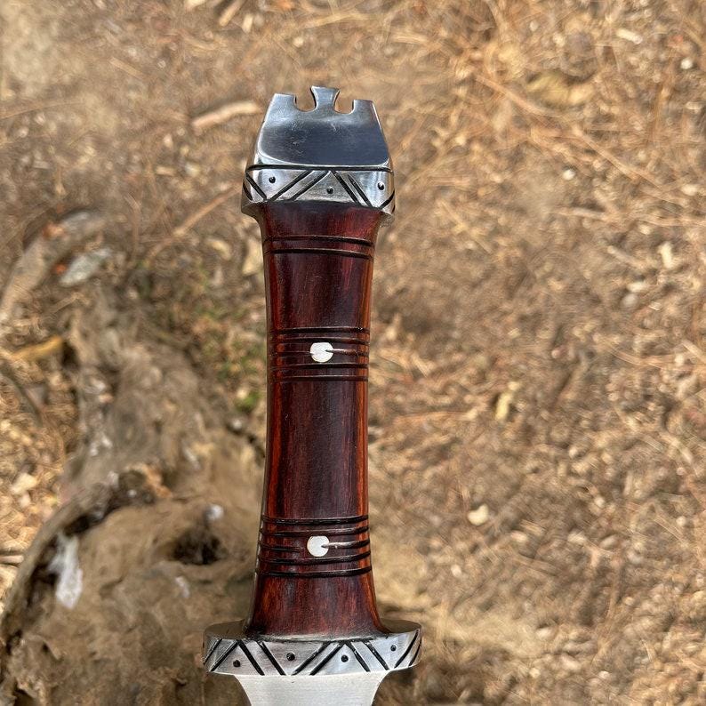 May include: A handcrafted knife with a dark wood handle and a silver-colored metal blade. The handle features decorative silver accents and a unique shape. The blade is partially visible, showcasing a sharp edge. The knife is set against a natural, outdoor background.