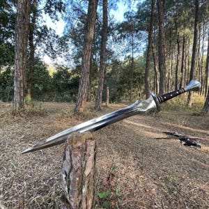 Puede incluir: Una espada plateada con un mango marr&oacute;n y negro descansa sobre un toc&oacute;n en un entorno forestal. La espada tiene una hoja larga y puntiaguda y una empu&ntilde;adura decorativa. El fondo presenta &aacute;rboles altos y hierba seca.