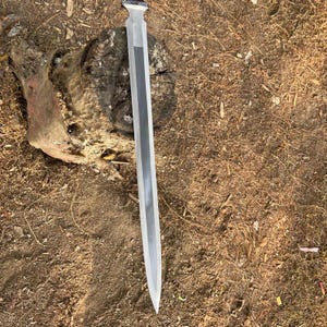 May include: A long, silver sword with a decorative handle. The blade has a central dark stripe and tapers to a sharp point. The sword is lying on a bed of brown leaves and dirt.