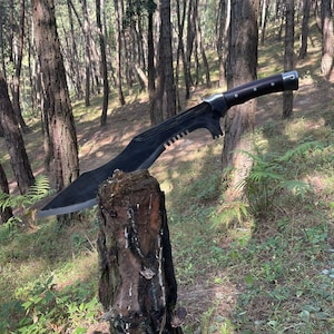 May include: A black machete with a wooden handle and a serrated edge. The machete is resting on a tree stump in a forest setting.