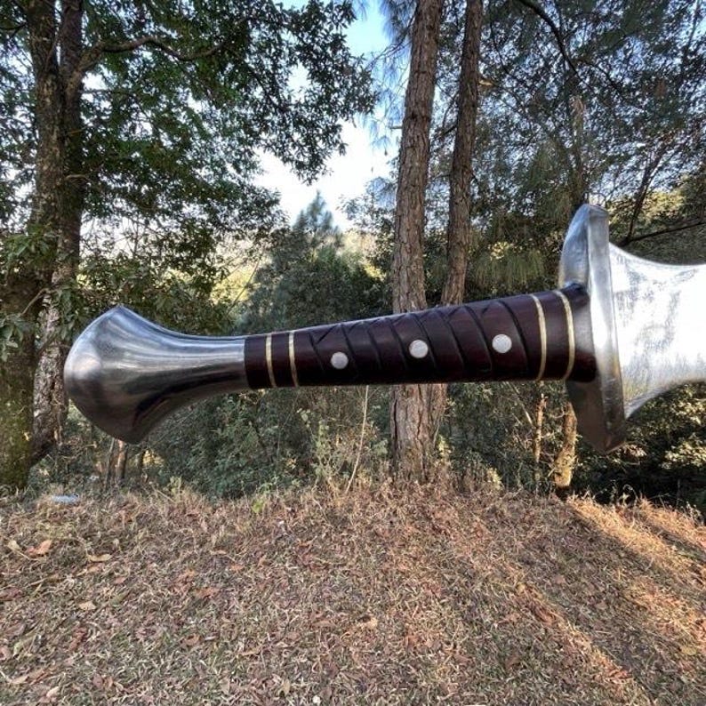 Puede incluir: Una espada de estilo medieval con una hoja de acero pulido y un mango envuelto en cuero marr&oacute;n oscuro. El mango presenta detalles en lat&oacute;n y tres remaches blancos. El pomo es de forma redondeada en acero pulido.