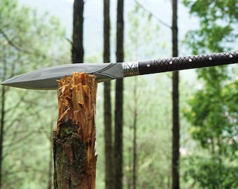 May include: A silver blade with a brown wooden handle is stuck into a tree stump. The blade is sharp and has a pointed tip. The handle is decorated with a pattern of small diamonds.