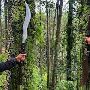 May include: Two silver swords with black handles are held by two people in a forest setting. The swords have a curved blade and a decorative design on the handle.