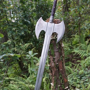 Könnte beinhalten: Ein silbernes Doppelkopf-Axt-Schwert mit einem Holzgriff, vor einem Hintergrund aus grünem Laub.