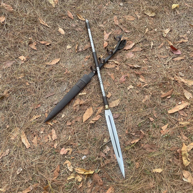 May include: A silver and black spear with a leather sheath. The spear is lying on a bed of brown leaves.