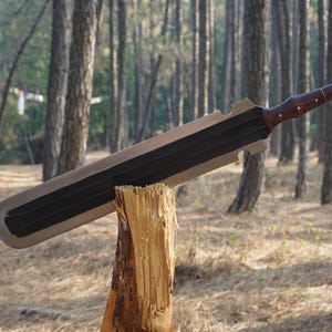 May include: A long, silver-colored sword with a black handle and a wooden grip. The sword is resting on a wooden stump in a forest setting.