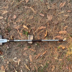 May include: A long, silver and brown spear with a pointed tip. The spear is lying on a bed of dry leaves.