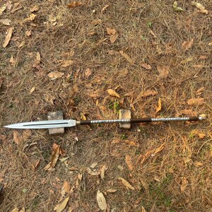 May include: A silver and brown spear with a wooden handle and a metal tip. The spear is lying on a bed of dry leaves.