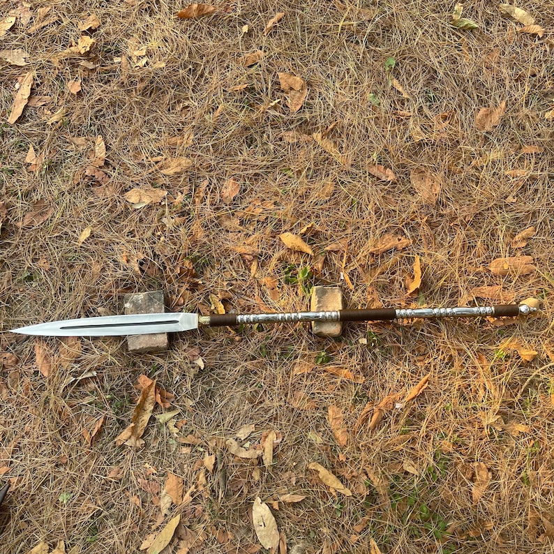 May include: A silver and brown spear with a wooden handle and a metal tip. The spear is lying on a bed of dry leaves.