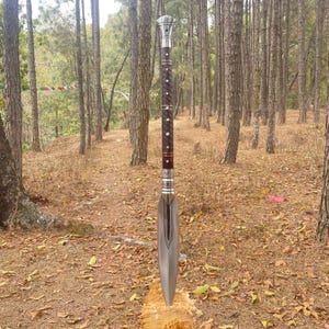 May include: A silver and brown spear with a wooden handle and a sharp point. The spear is standing upright in a forest setting.