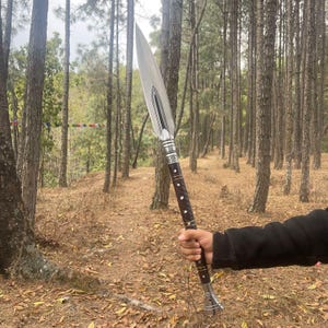 May include: A silver spear with a wooden handle and a decorative metal tip. The spear is held by a person in a forest setting.