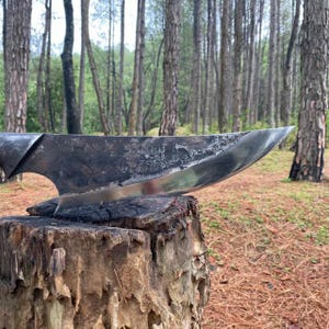 May include: A large, hand-forged knife with a curved blade, resting on a weathered tree stump. The blade has a dark, textured finish with a polished edge. The background features a forest with tall trees.
