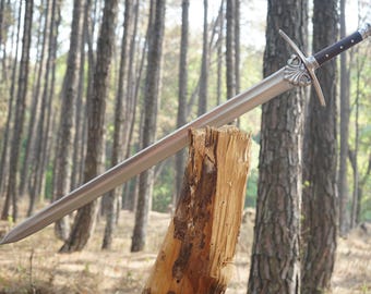 May include: A silver sword with a black handle and a decorative pommel is propped against a tree stump in a forest setting.