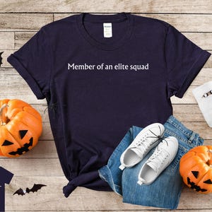 May include: A navy blue t-shirt with the text "Member of an elite squad" printed in white. The shirt is laying on a wooden surface with a pair of white sneakers, blue jeans, and a pumpkin.