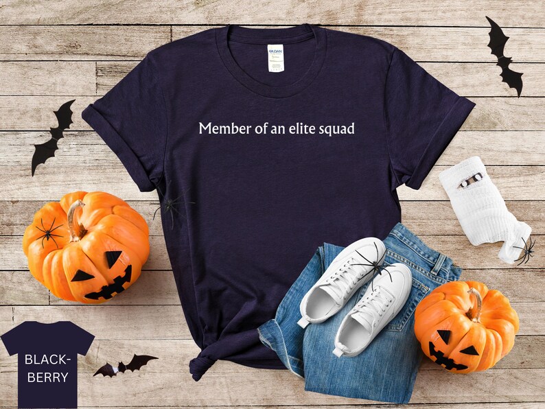 May include: A navy blue t-shirt with the text "Member of an elite squad" printed in white. The shirt is laying on a wooden surface with a pair of white sneakers, blue jeans, and a pumpkin.