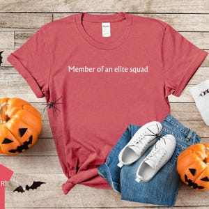 May include: A red heather t-shirt with the text "Member of an elite squad" printed in white on the front. The shirt is laid out on a wooden surface with a pair of white sneakers, blue jeans, and a white sock with a black spider on it.